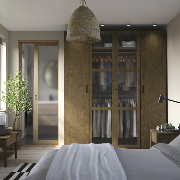 Chambre avec grande armoire PAX en bois clair, suspension suspendue, lit blanc avec couverture, petit bureau avec lampe et arbre en pot à côté de la porte.