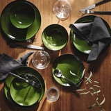 Set de vaisselle, vert foncé, brillant. Assiettes, bols, verres, couverts disposés sur une table en bois.