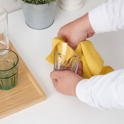 Personne essuyant des verres avec un chiffon jaune sur une table blanche. Deux verres, une plante et une planche de bois à proximité.