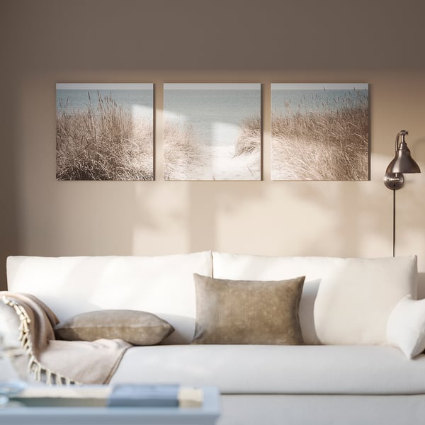 Trois photos de plage dans des cadres blancs sur un mur marron. Canapé blanc avec oreillers, couverture et lampe argentée à côté.