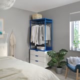 Chambre moderne avec des murs gris, un lit blanc, meuble de rangement PLATSA bleu. Des étagères contiennent des vêtements, des paniers et une plante. Une chaise et une fenêtre avec des stores.