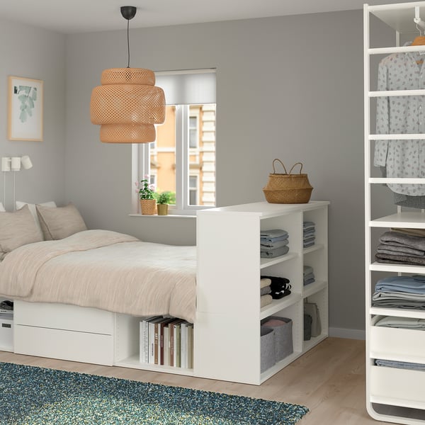 Chambre minimaliste avec lit PLATSA, rangements et livres. Parquet clair, murs gris, tapis fleuri. Grande fenêtre, suspension chaleureuse.