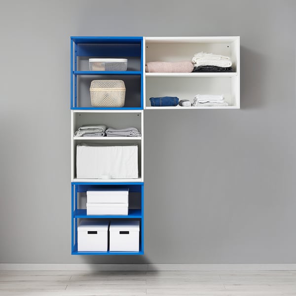 Une unité de rangement modulaire bleue et blanche, PLATSA, montée sur un mur, avec des vêtements soigneusement pliés et des boîtes de rangement sur des étagères.