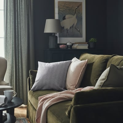 Salon confortable avec canapé vert, coussins rayés bleu/blanc, lampe et armoire noire avec de curieuses œuvres d’art de grue.