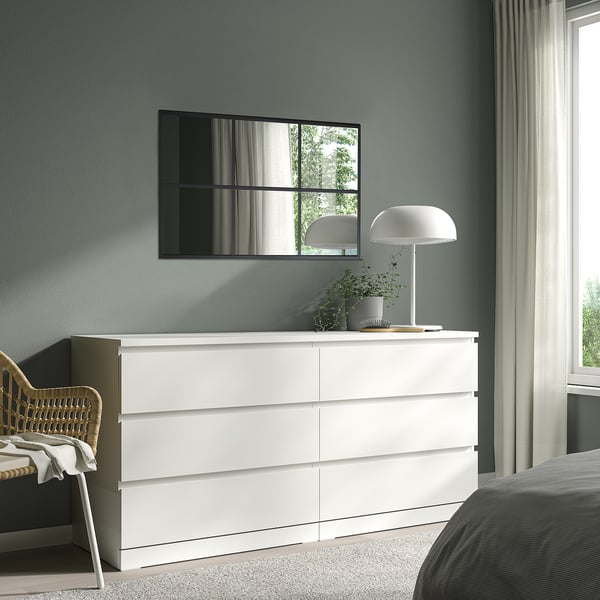 Miroir RAGGARÖN noir au-dessus d’une commode blanche, fenêtre réfléchissante et arbres. Commode à six tiroirs, chaise en osier, lit gris avec literie rayée.