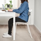 Personne assise à un bureau à l’aide d’un ordinateur portable, portant une chemise en jean, un pantalon noir et des baskets blanches, avec un coussin de chaise blanc offrant un soutien lombaire.