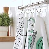 Une rangée de torchons de cuisine colorés sont suspendus à des crochets, avec des motifs de carrés, de points et d’ustensiles de cuisine, parfaits pour nettoyer et sécher la vaisselle.
