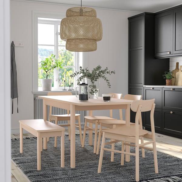 Salle à manger à la lumière naturelle avec table RÖNNINGE en bois, chaises assorties, banc, verdure, lampe en bambou, armoires sombres et tapis neutre.