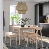 Salle à manger à la lumière naturelle avec table RÖNNINGE en bois, chaises assorties, banc, verdure, lampe en bambou, armoires sombres et tapis neutre.