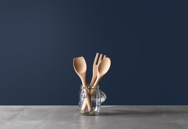 Quatre ustensiles en bois dans un bocal transparent sur une surface grise, mur bleu.