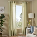 Des rideaux transparents vert olive sont suspendus à une fenêtre, complétant les murs beiges et un canapé blanc. Une plante en pot et une lampe latérale sont également présentes.