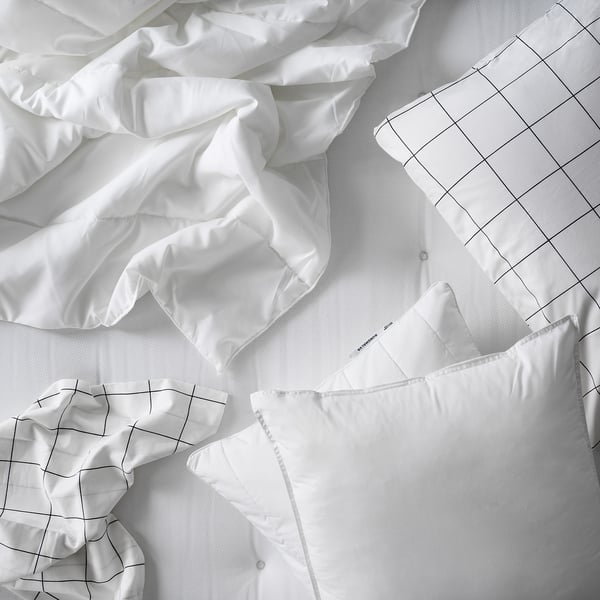 Une couette SÄFFEROT blanche. Il est étalé sur un lit avec un drap à motifs quadrillés. La couette est douce et moelleuse, remplie de polyester recyclé.