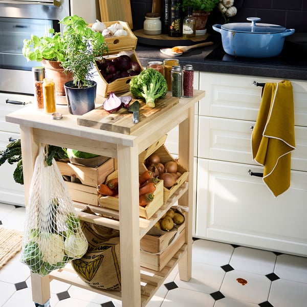 Îlot de cuisine en bois avec des caisses de légumes, des herbes dans des pots et des ustensiles de cuisine sur le dessus. Une serviette jaune pend à une poignée de tiroir.