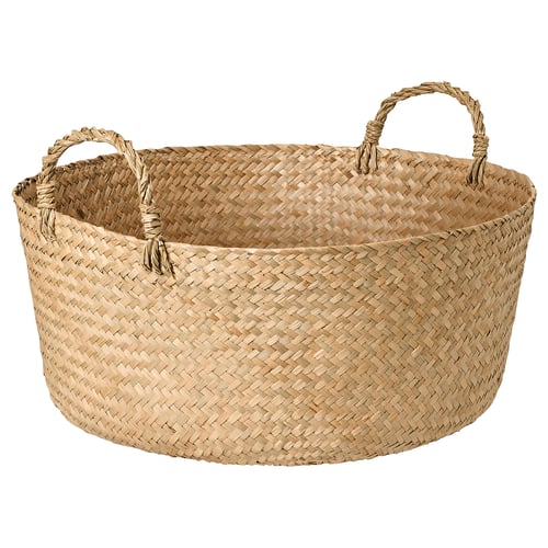 Un panier tressé SANDJORD fait à la main. Il est beige, circulaire et fabriqué à partir d’un matériau de carex. Il est doté de deux poignées pour un transport facile.