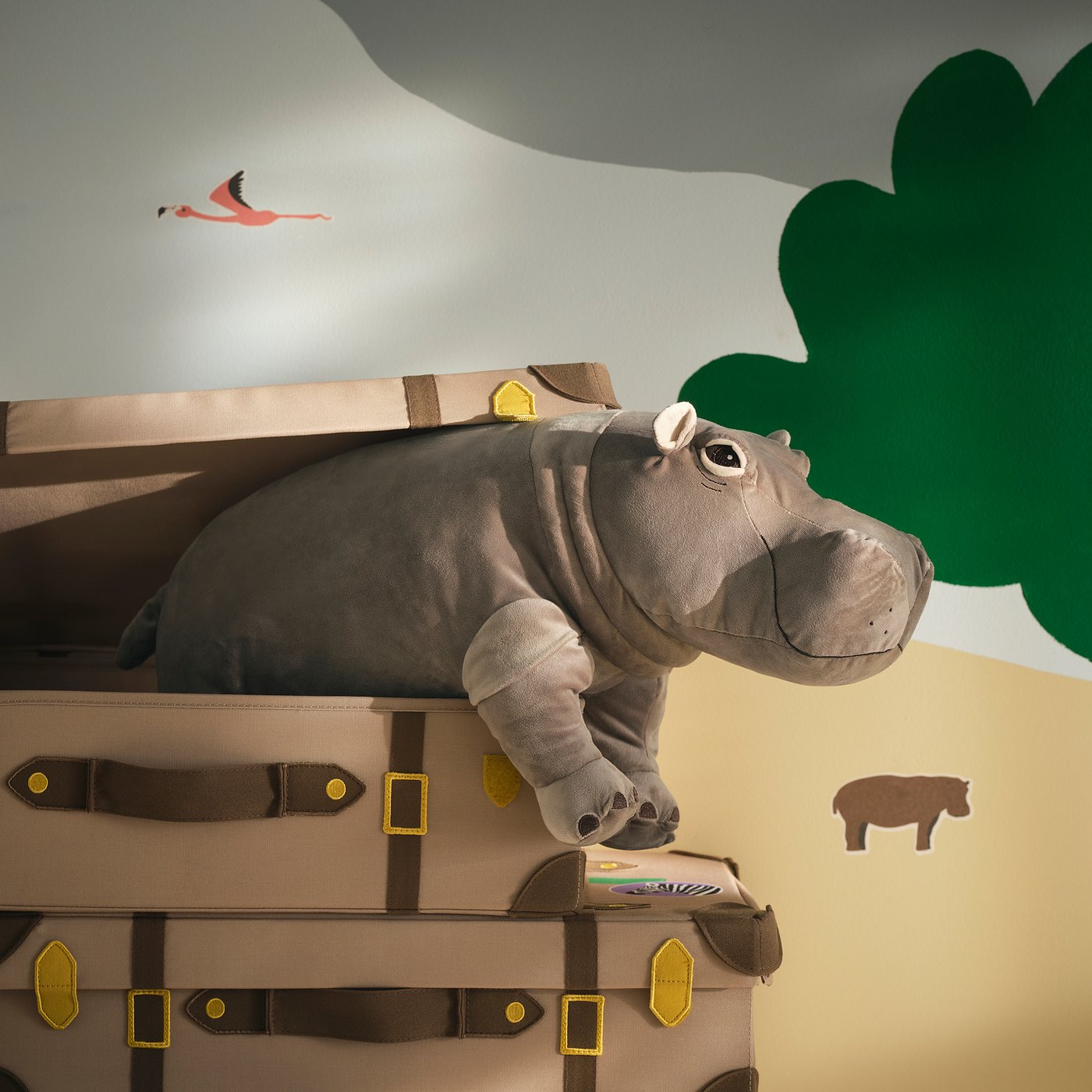 Un jouet hippopotame gris en peluche est posé sur de grands coffres de voyage beiges, plus grands que les coffres eux-mêmes, avec une étiquette jaune.