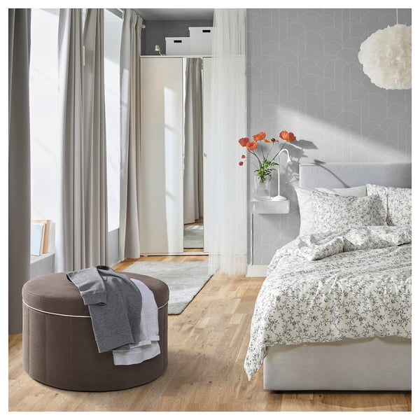 Chambre lumineuse avec des murs gris clair, un grand lit, une literie florale et un pouf rond marron avec une couverture drapée dessus.