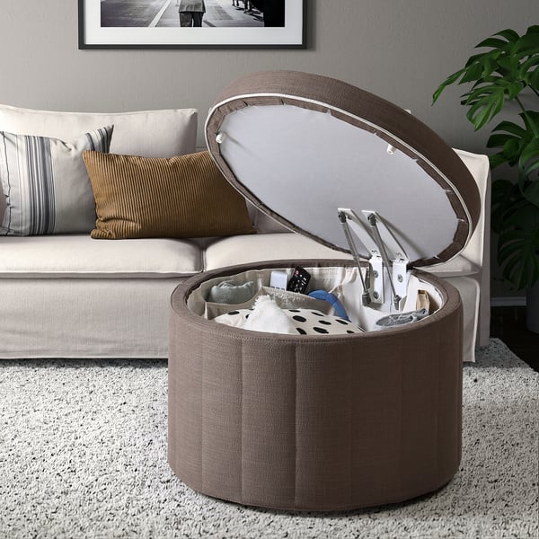 Pouf de rangement rond ouvert, révélant divers objets dans les poches. Placé à côté d’un canapé avec des oreillers, sur un tapis blanc moucheté.