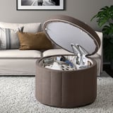 Pouf de rangement rond ouvert, révélant divers objets dans les poches. Placé à côté d’un canapé avec des oreillers, sur un tapis blanc moucheté.