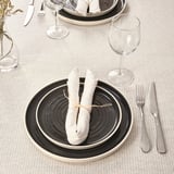 Dressage de table avec des assiettes noires et blanches, des serviettes roulées, de l’argenterie et des verres à vin sur une nappe beige.