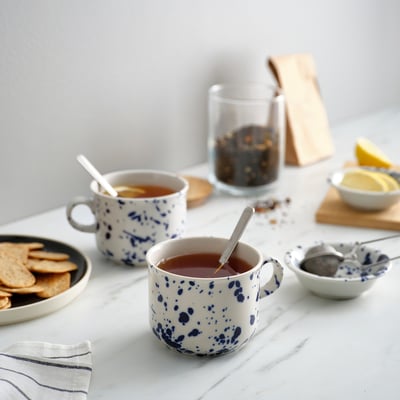 Deux mugs blancs SILVERSIDA à pois bleus. L’un tient du thé, une cuillère à l’intérieur. Plus petit que les assiettes avec des craquelins et des tranches de citron. Fabriqué en grès.