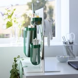 Espace de travail organisé avec un panneau perforé SKÅDIS, des poches en filet vert contenant des fournitures de bureau et des conteneurs blancs.