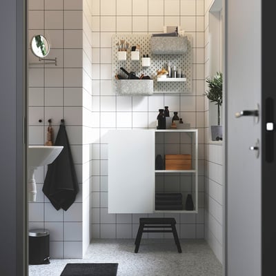 Un petit panier SKÅDIS noir est suspendu à un panneau perforé, rempli d’objets, positionné près d’un lavabo et d’une armoire de salle de bain.
