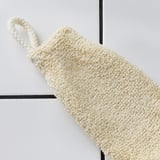 Une mitaine exfoliante de couleur crème est suspendue à un crochet, montrant sa surface de gommage texturée et douce, parfaite pour le bain.