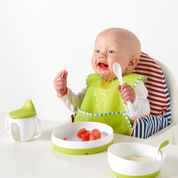 Bébé dans une chaise haute mange dans une assiette à bords inclinés, gardant la nourriture à l’intérieur. Bavoir vert et gobelet à bec à côté.