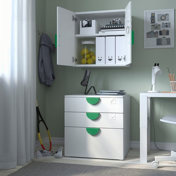 Unité de rangement blanche avec armoire ouverte exposant jouets et livres. Poignées et crochets verts avec un bureau à proximité et des équipements de sport.