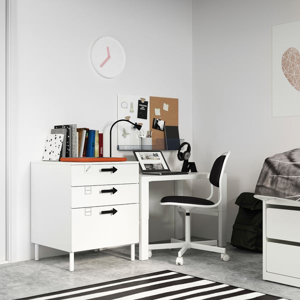 Chambre d’enfant moderne avec coffre et bureau SMÅSTAD blancs, chaise noire, décor coloré, tapis rayé et grande horloge.