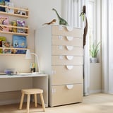 Chambre d’enfant avec commode beige SMÅSTAD, tiroirs, bureau, lampe, tabourets, jouets dinosaures, livres et plante.