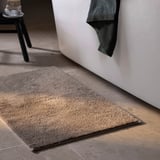 Un tapis de bain gris SÖDERSJÖN se trouve près d’une baignoire blanche. Fabriqué en 100 % coton, il est composé d’un poil épais et doux et d’un support en latex pour plus de stabilité.
