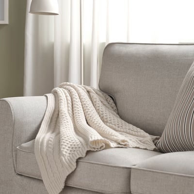 un plaid crème SÖRBYSKOGEN drapé sur un canapé gris ; doux, confortable, de taille moyenne.