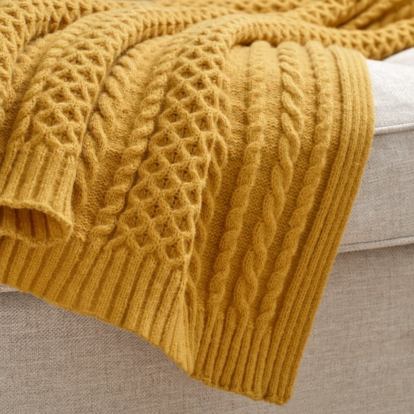 Une couverture en tricot jaune moutarde douillette aux motifs complexes, drapée sur un canapé beige.