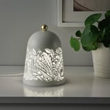 Une lampe de table décorative, SOLSKUR, avec un design inspiré de la nature, offre un éclairage d’ambiance doux, assis sur une surface blanche à côté d’une plante en pot.