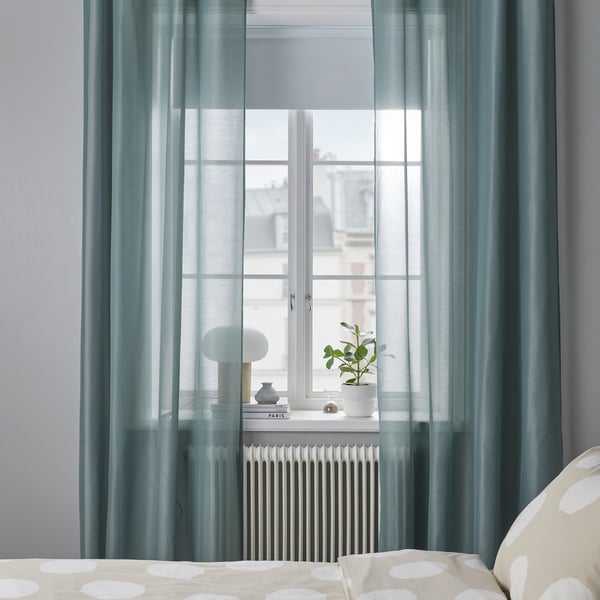 Des rideaux transparents turquoise STENFRÖ avec œillets couvrent la grande fenêtre, atténuant la lumière et ajoutant une ambiance côtière. Lit et plante visibles.