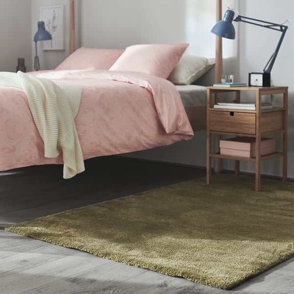 Chambre moderne avec tapis moelleux vert olive STOENSE. Linge de lit rose, table de chevet en bois, lampe et éparpillement de livres.