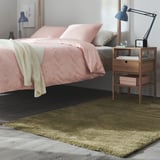Chambre moderne avec tapis moelleux vert olive STOENSE. Linge de lit rose, table de chevet en bois, lampe et éparpillement de livres.