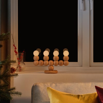 La lampe STRÅLA brille doucement sur le rebord de la fenêtre avec un décor de vacances, en bois et sphérique, à côté d’un coussin jaune.