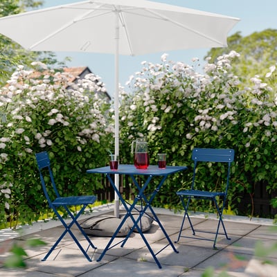 Ensemble de patio en métal bleu avec table et chaises sous un parasol blanc, situé dans un jardin luxuriant.