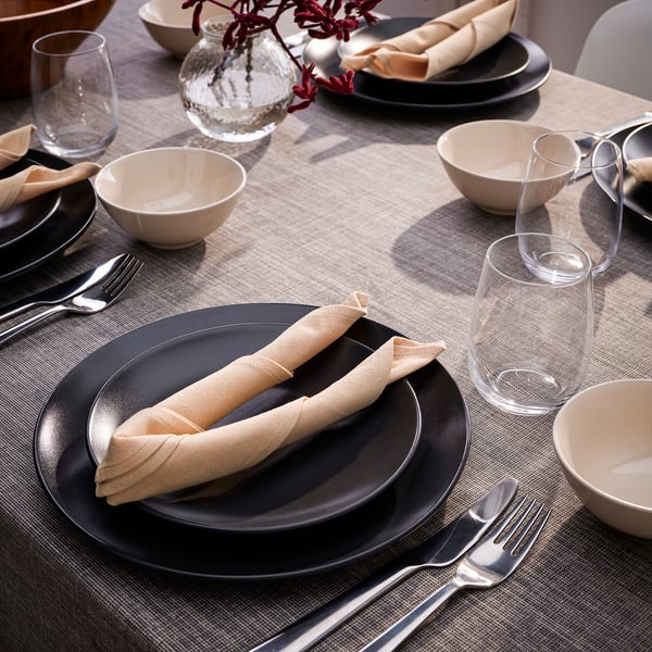 Set de table avec des assiettes noires, des bols beiges, des serviettes beiges roulées, des verres transparents et de l’argenterie, soigneusement disposés sur une nappe beige.