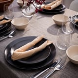 Set de table avec des assiettes noires, des bols beiges, des serviettes beiges roulées, des verres transparents et de l’argenterie, soigneusement disposés sur une nappe beige.