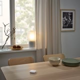 Salle à manger avec télécommande SYMFONISK, lampe, pommes de pin et plats empilés. Chaises minimalistes et art des papillons.