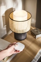 Une main tient une télécommande blanche SYMFONISK près d’une table avec une lampe tissée noire et beige.
