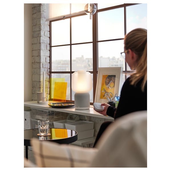 La femme travaille par une fenêtre lumineuse dans une pièce simple avec bureau et lampe.