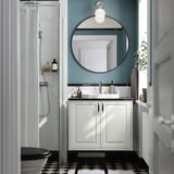 Salle de bains moderne TÄNNFORSEN vanité blanche, miroir rond, carrelage noir et douche en verre. Lavabo et toilettes visibles, avec des murs bleus.