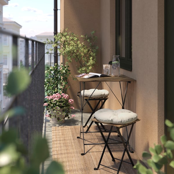 De petits tabourets en bois et métal noir avec des coussins sur un balcon, à côté de plantes en pot et d’une table avec un livre et des boissons.