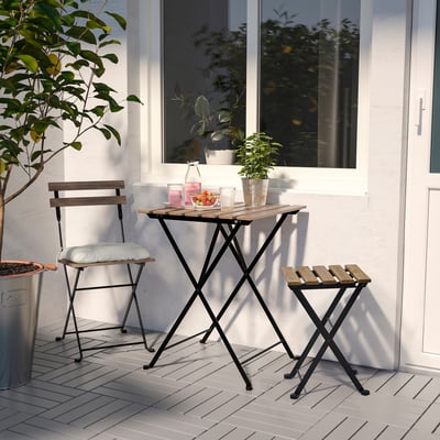 Petite installation extérieure avec table, chaise et tabouret pliants TÄRNÖ en bois naturel et métal noir. Parfait pour les espaces compacts, entourés de plantes.