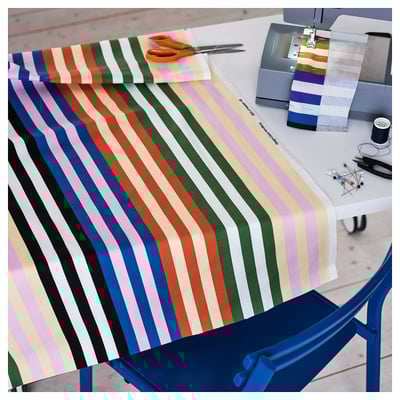 Tissu rayé brillant sous machine à coudre sur table. Des ciseaux orange et une bobine de fil sont à proximité, indiquant la préparation du projet de couture.