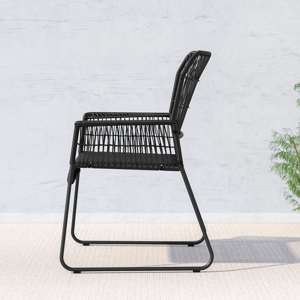 Chaise d’extérieur noire en rotin tressé, conçue pour le confort et la durabilité, positionnée contre un mur clair avec une plante.